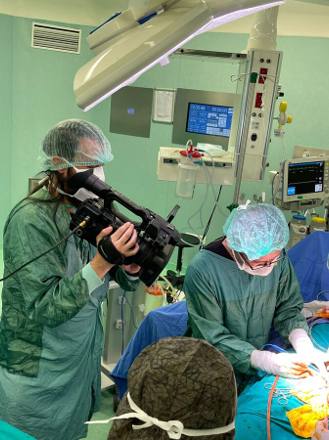 A camera operator in a gown and cap filming over the shoulder of the operating surgeon, with the theatre clock and patient monitor visible behind.