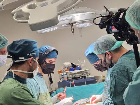 A broadcast video camera on a rig recording the operating table while the gowned team works beneath the theatre lamp array.