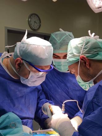 The gowned team at the operating table during an implant procedure, working under the theatre lamp with the wall clock behind them.
