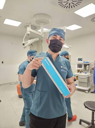 A team member in scrubs holding the sealed carton of a Coloplast penile implant in a bright modern operating room before the case begins.