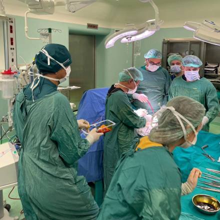 A wide view of the operating room with the gowned team at the table and a scrub nurse laying out clamps and forceps on the instrument trolley.
