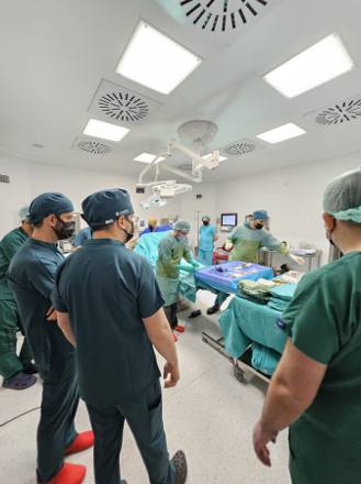 The operating room from the doorway, ceiling lamps and columns overhead, with the theatre team assembled around the table.