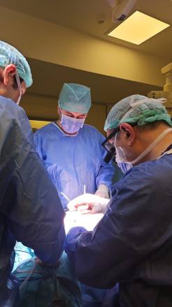 Two surgeons in blue gowns working over the draped operative field under a focused theatre light, the rest of the room in shadow.