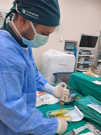 A scrubbed surgeon in a Coloplast Interventional Urology cap preparing an inflatable penile prosthesis at the back table, drawing solution into a syringe beside sterile bowls and instruments.