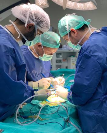 Four scrubbed team members in blue gowns and caps gathered over the sterile drapes beneath twin LED theatre lamps.