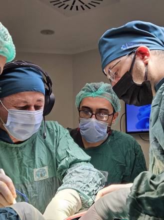 Three surgeons at the operating table in green gowns and caps, the operator wearing a headband loupe while two assistants watch the field.