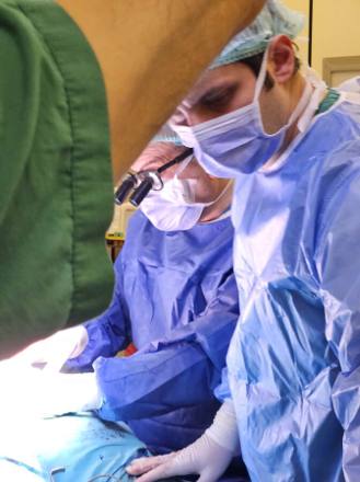 A close side view of a gowned surgeon working at the operating table, a colleague’s shoulder in the foreground.