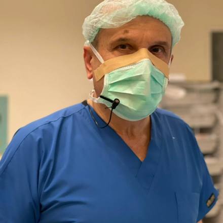 A senior surgeon in blue scrubs, surgical cap and mask standing in the operating room with a clip microphone on the collar during a filmed case.