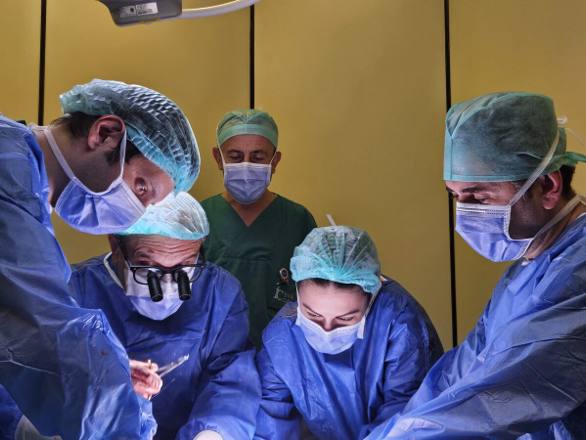 Masked and capped theatre staff in gowns leaning in around the operating table together during a procedure.