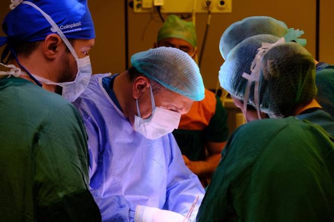 Surgeons in green and blue gowns bent over the illuminated operative field in a dimmed operating room.