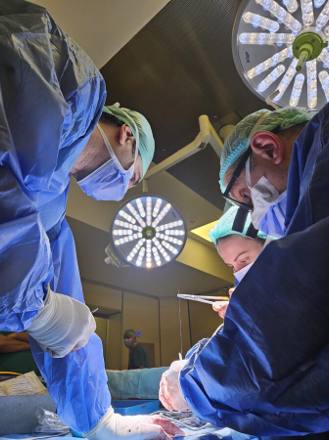 A large multi-lamp theatre light directly above the surgical team, who are working at the draped table in blue gowns.