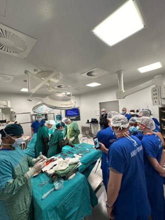 A wide view of a bright, modern operating room with the full team standing around the table and the instrument trolley alongside.