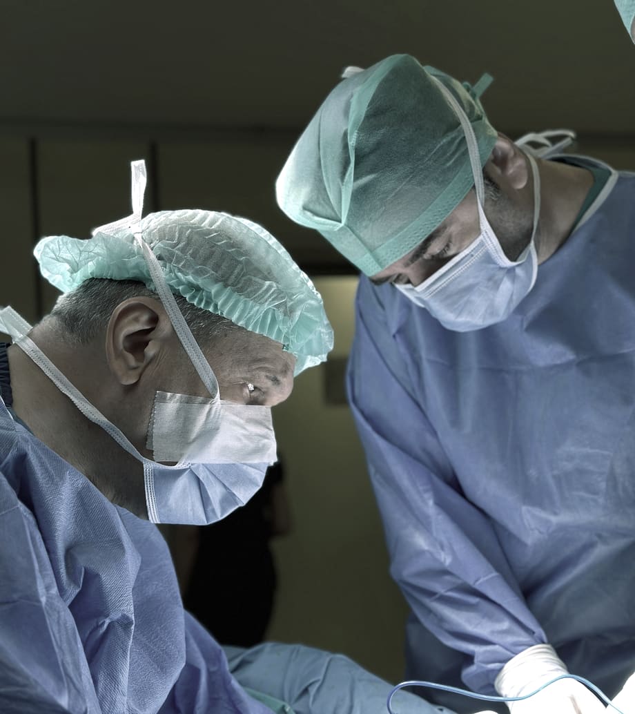 Two gowned and masked members of a surgical team working head-down over the operating table under theatre lights.