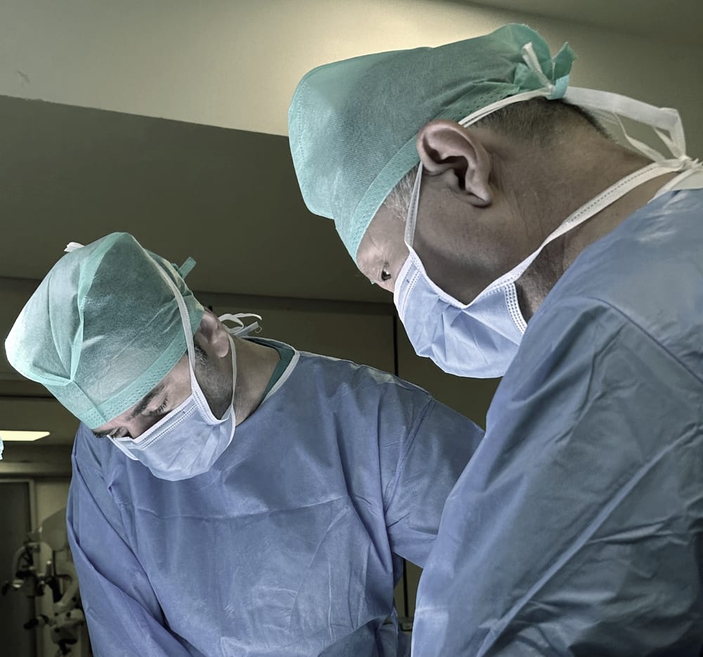 Two scrubbed and masked members of a surgical team, gowned and gloved, working head-down at the operating table.
