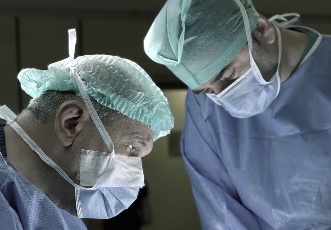 Two masked surgeons in gowns and caps, heads down and working at close quarters in an operating theatre.